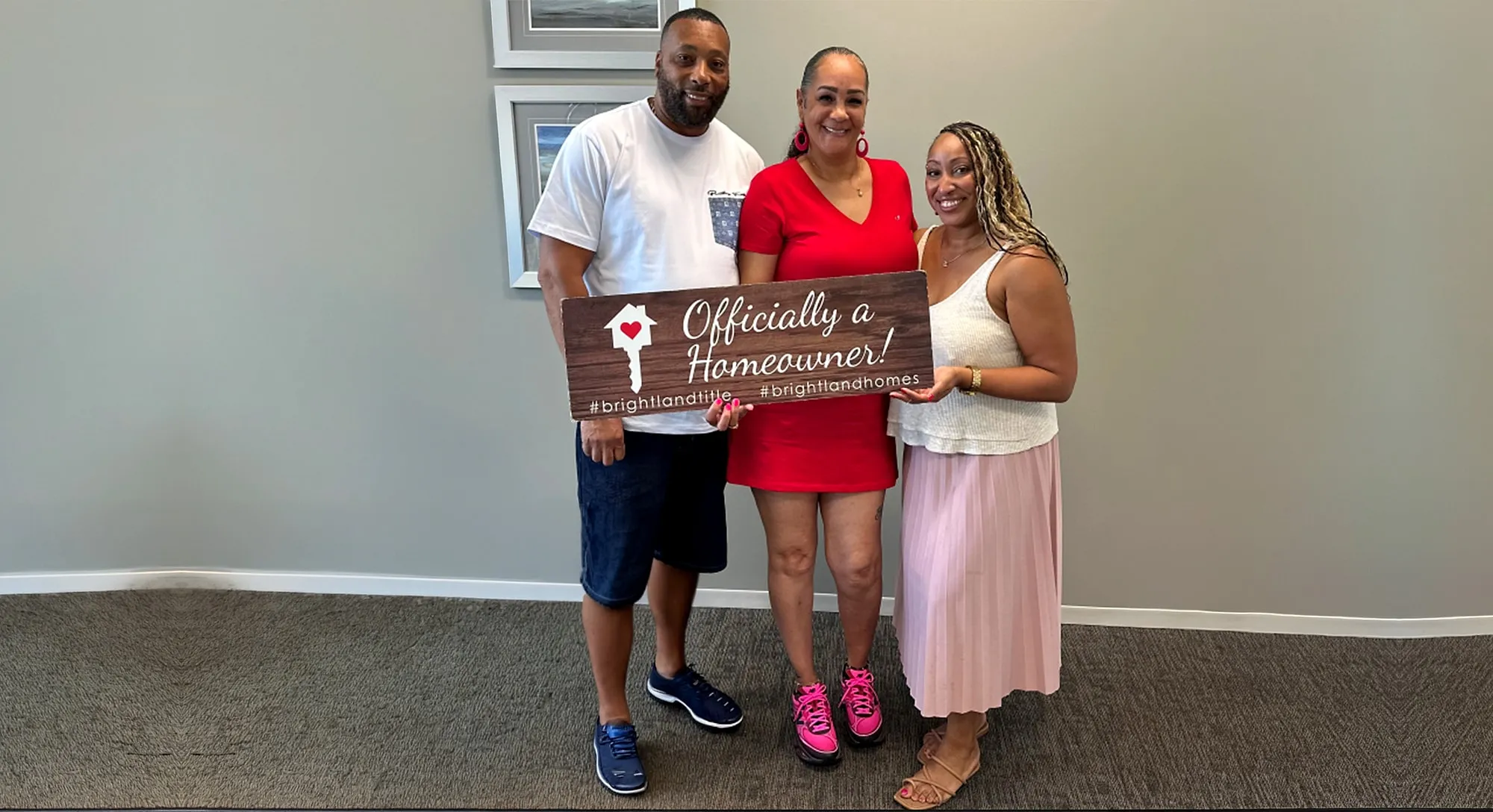 Three people standing in an office holding a wooden sign that reads “Officially a Homeowner!”
