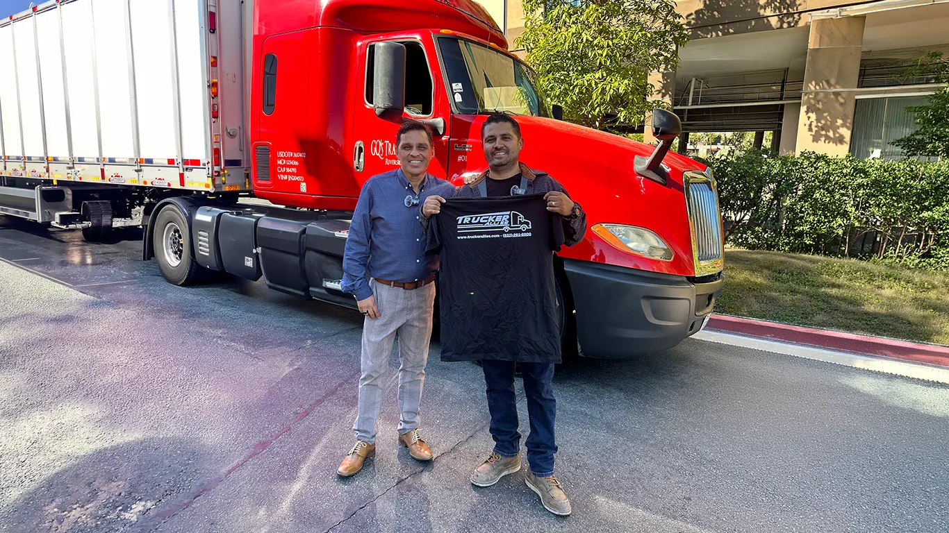 Two men standing in front of a red GQ’s Transport semi truck, one holding up a Trucker Allies shirt.
