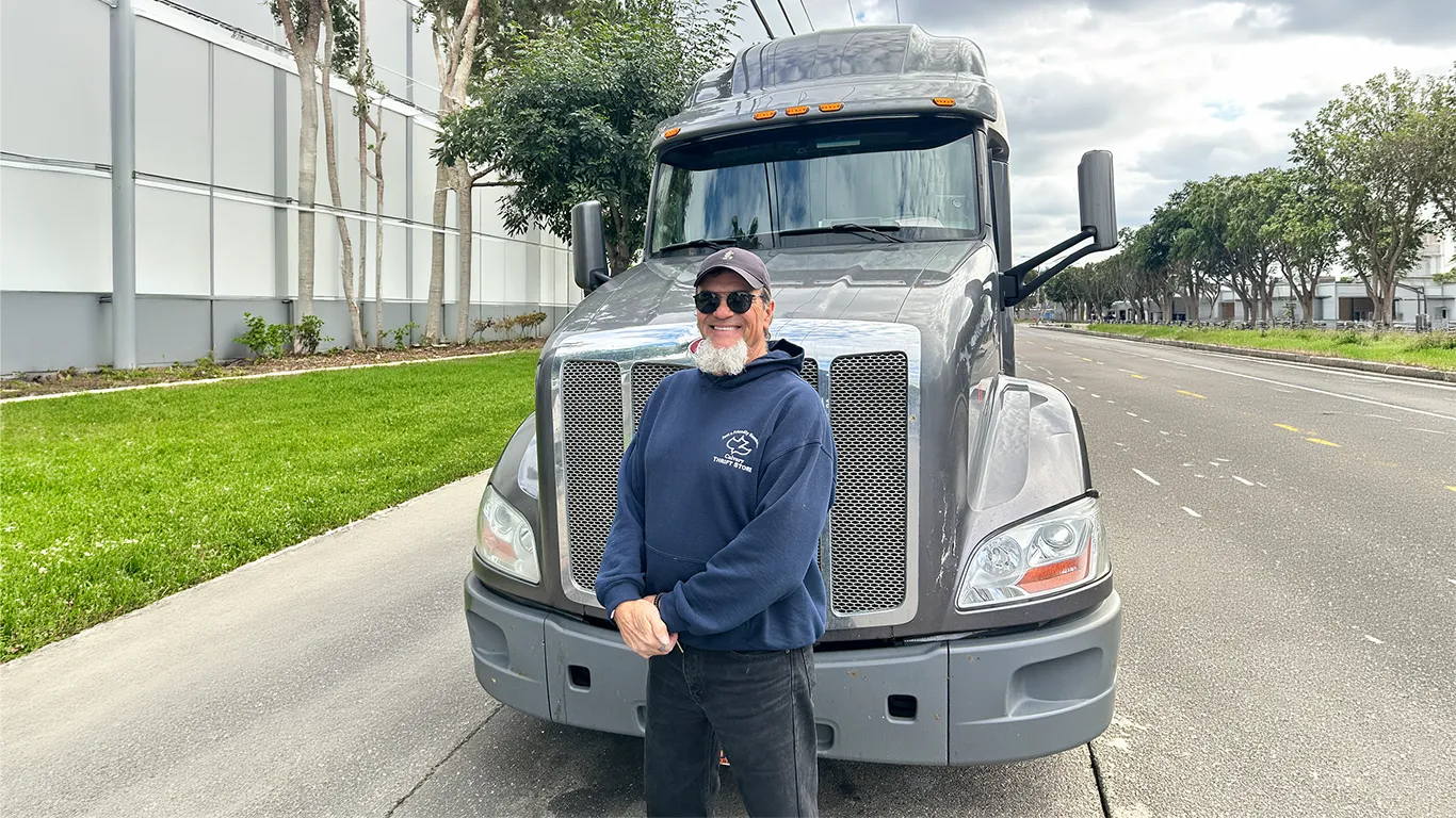 A truck driver in sunglasses and a navy hoodie standing in front of the grille of a grey semi truck.